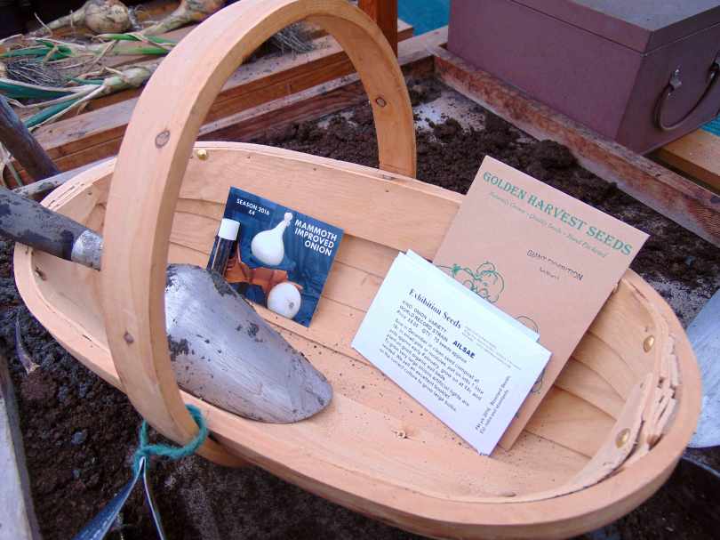Picture of a trowel and packets of giant onion seeds in a wooden trug