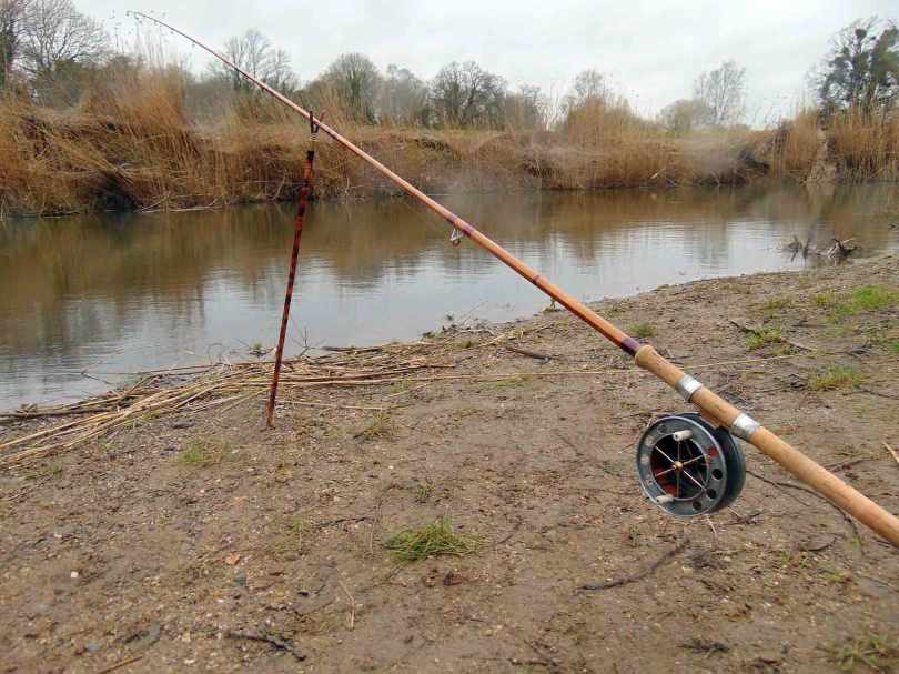 Picture of a Fred J. Taylor rod and Allcocks Aerial fishing reel sitting in a rod rest at the edge of a river.